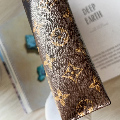 Brown leather wallet with gold monogram pattern on a surface with books and a pen.