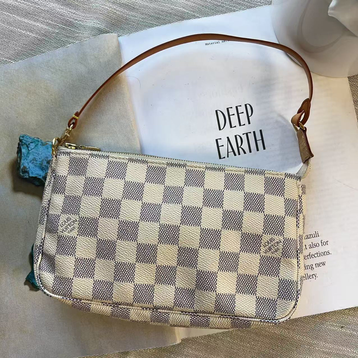 A small checkered shoulder bag with a brown leather strap, resting on an open book with the words 'DEEP EARTH' on the page, set on a textured gray fabric surface.