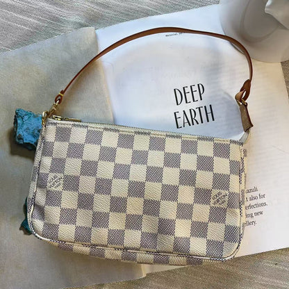 A small checkered shoulder bag with a brown leather strap, resting on an open book with the words 'DEEP EARTH' on the page, set on a textured gray fabric surface.