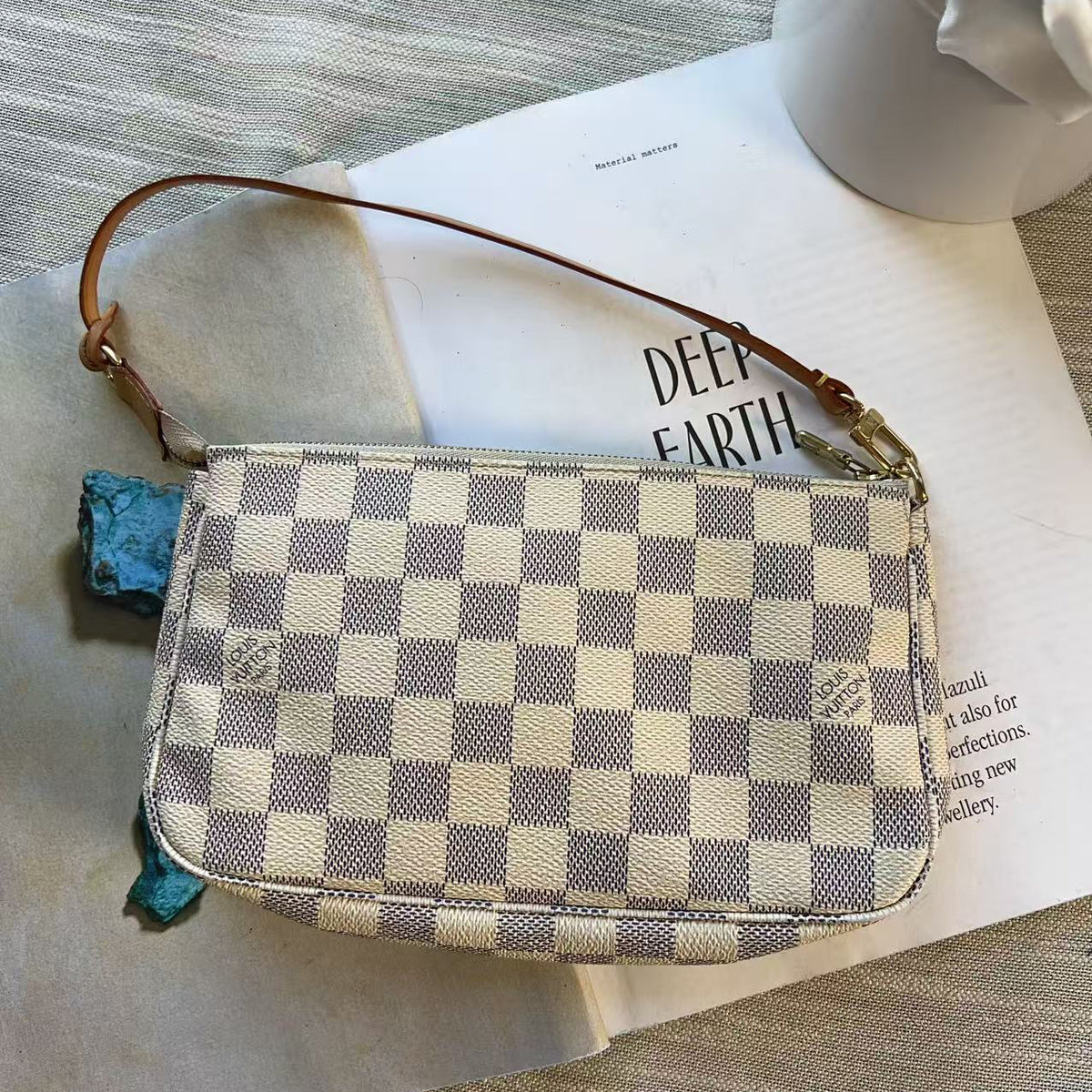 A Louis Vuitton shoulder bag in the Damier Azur checkerboard pattern, featuring a brown leather strap and gold-tone hardware, styled on top of a book and a beige fabric surface.