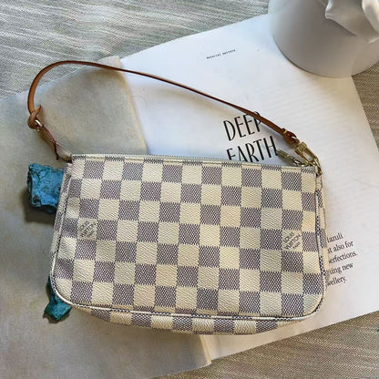 A Louis Vuitton shoulder bag in the Damier Azur checkerboard pattern, featuring a brown leather strap and gold-tone hardware, styled on top of a book and a beige fabric surface.