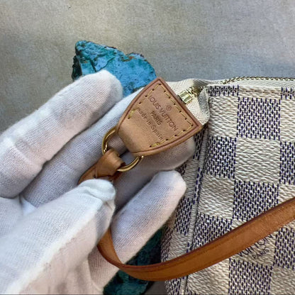 A close-up of a Louis Vuitton handbag with a beige and navy checkered pattern, featuring a tan leather trim and a gold-tone metal ring. A hand wearing a white glove holds the leather detail, set against a neutral textured background.