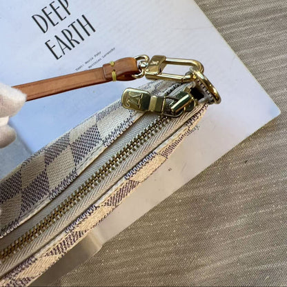 A close-up of a zippered pouch with a beige and gray plaid pattern, gold-tone hardware, and a brown leather pull tab, resting on a white book over a textured gray surface.