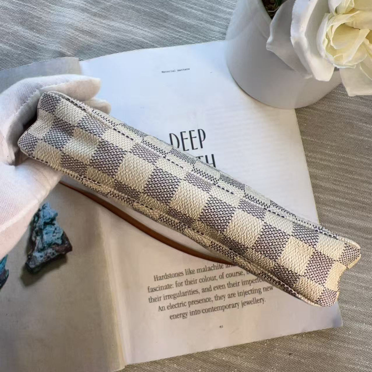 A checkered canvas pencil case with a beige and gray pattern, resting on an open book with a white glove holding one end, styled with a white flower pot in the background.
