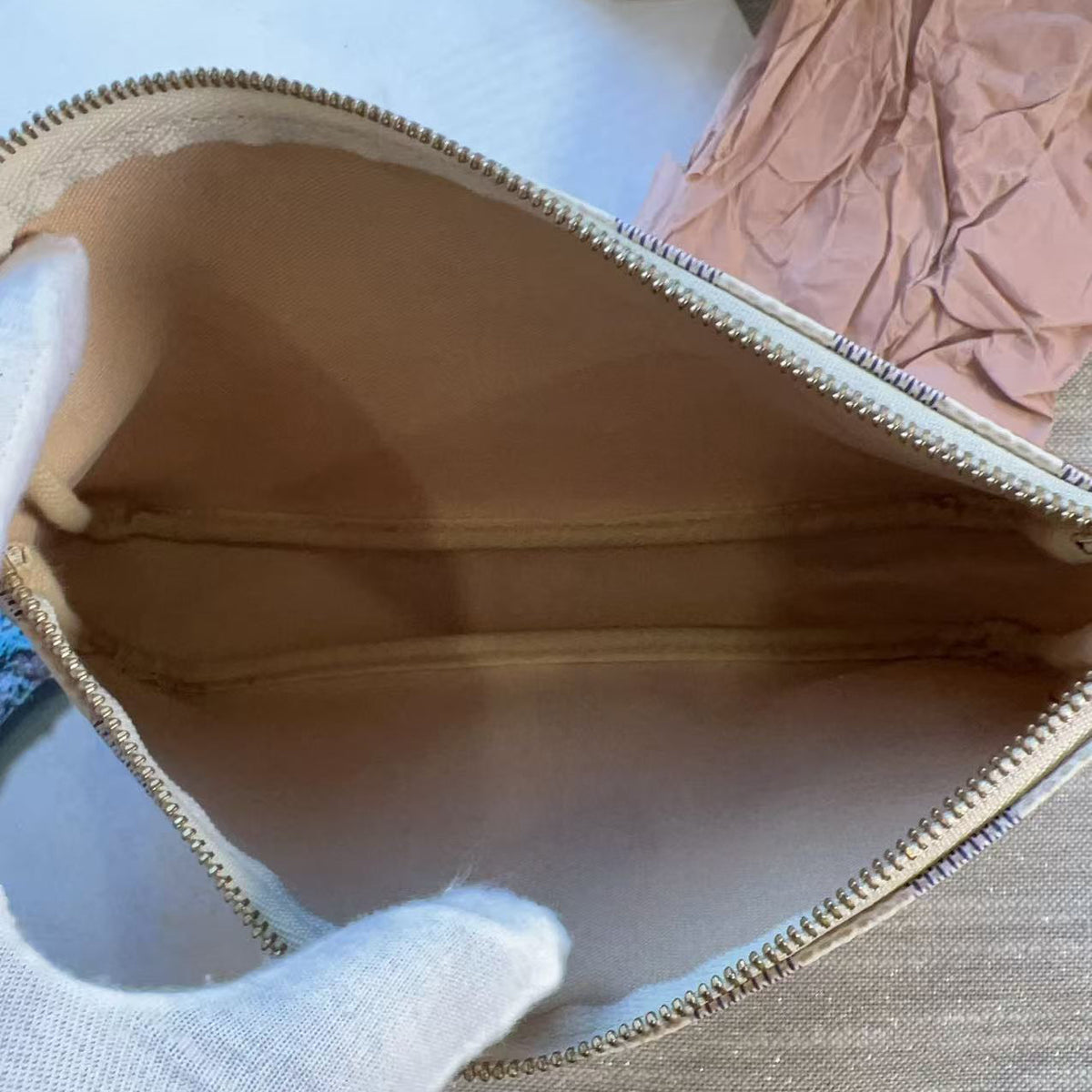 A close-up of a beige zippered pouch or cosmetic bag being held open by a white gloved hand, with a light-colored fabric surface and a pink crumpled paper visible in the background.