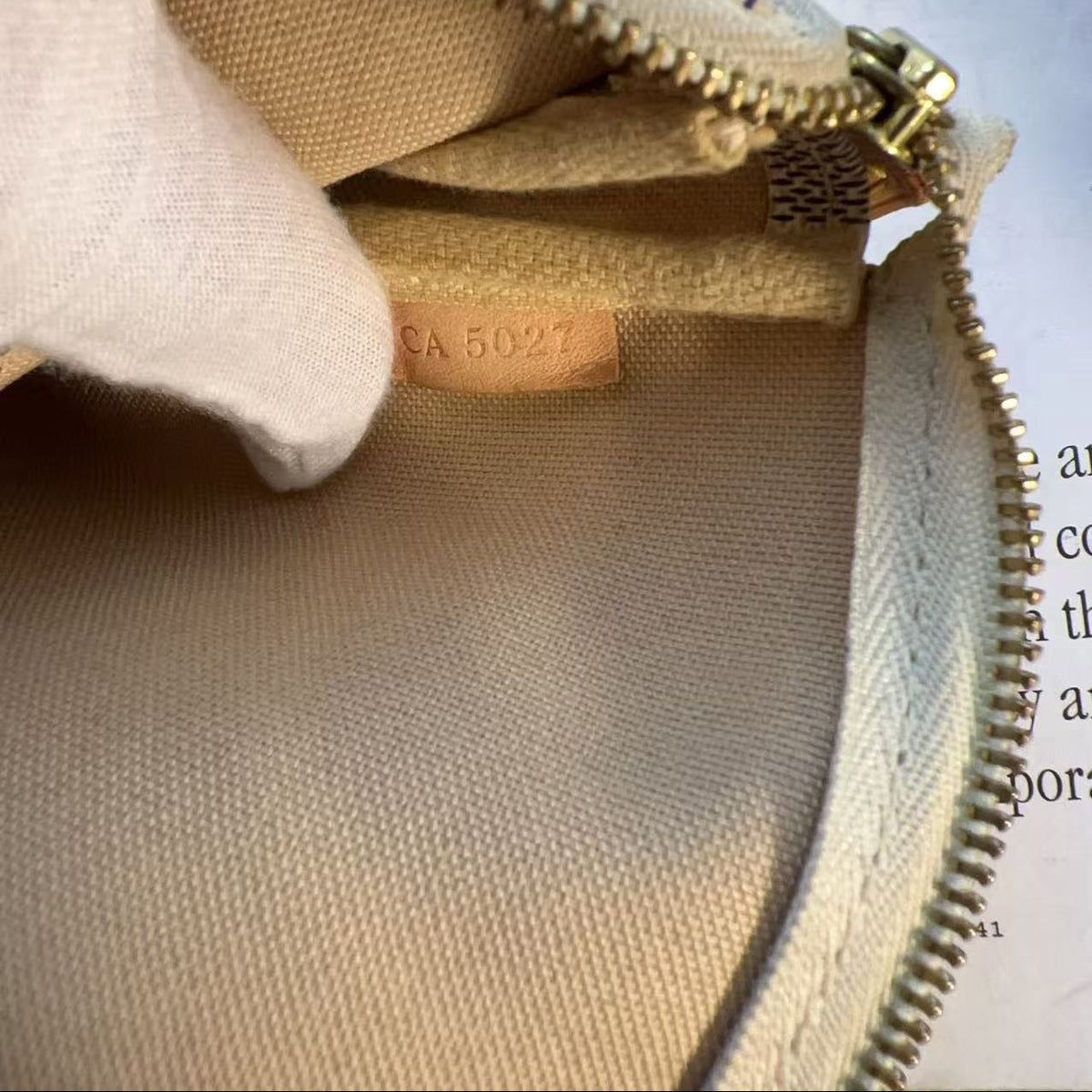 Close-up of a beige canvas zippered pouch or bag interior with a tan leather label reading 'CA 5027' and a white fabric pull tab, photographed against printed paper with partial text visible.
