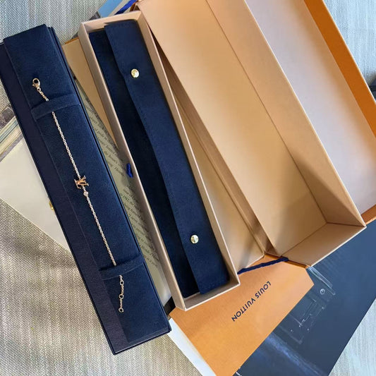 A gold-tone chain bracelet with a small interlocking LV logo pendant is displayed inside a navy blue velvet jewelry box, alongside other Louis Vuitton packaging boxes on a textured gray fabric background.