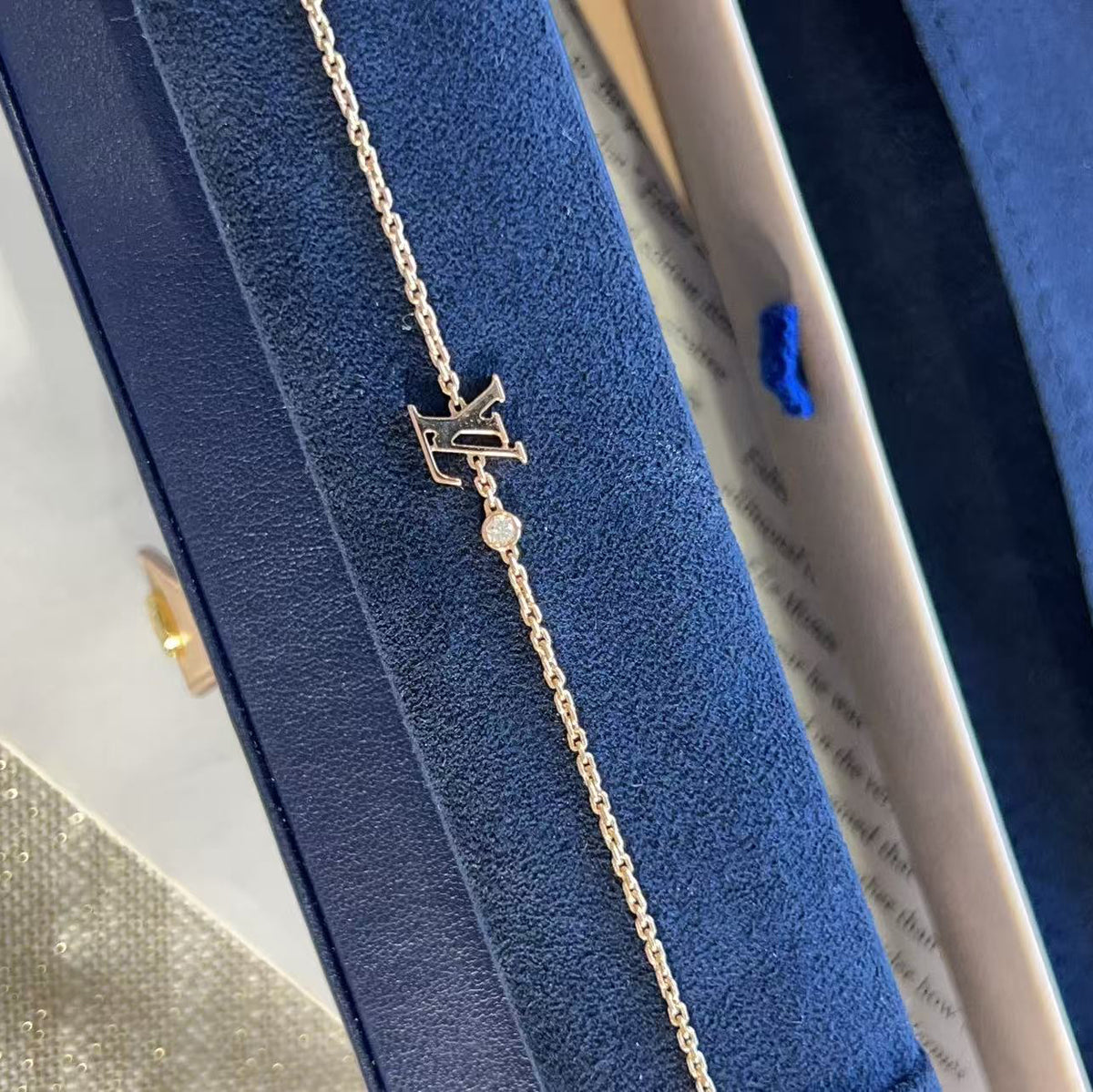 A delicate gold chain bracelet with a custom monogram pendant and a small clear stone, displayed on a textured navy blue jewelry box.