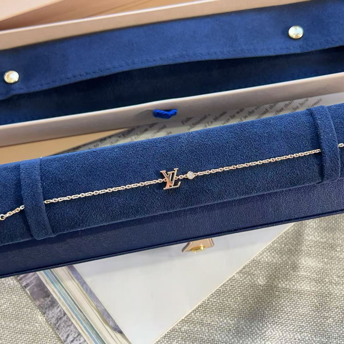 A delicate gold chain bracelet with a 'KA' monogram charm and a small clear stone, displayed inside a navy blue velvet jewelry box.