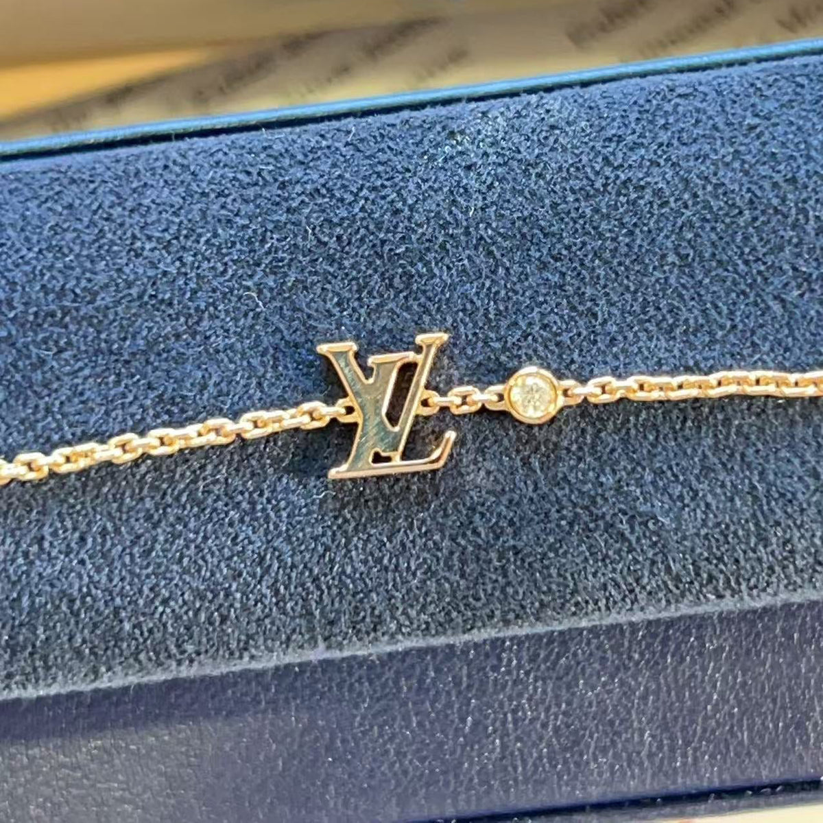 A gold-tone Louis Vuitton bracelet with an LV monogram charm and a small round clear stone, displayed on a blue velvet jewelry box.
