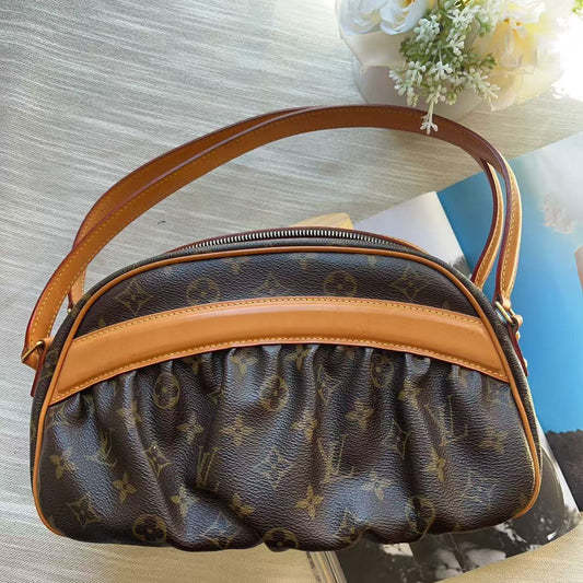 A Louis Vuitton monogram shoulder bag in brown coated canvas with tan leather trim and handle, resting on a light wood surface next to a book and white flowers.
