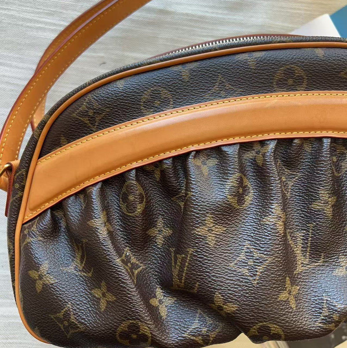 A close-up of a Louis Vuitton monogram canvas bag with tan leather trim and a matching shoulder strap, resting on a light wood surface.
