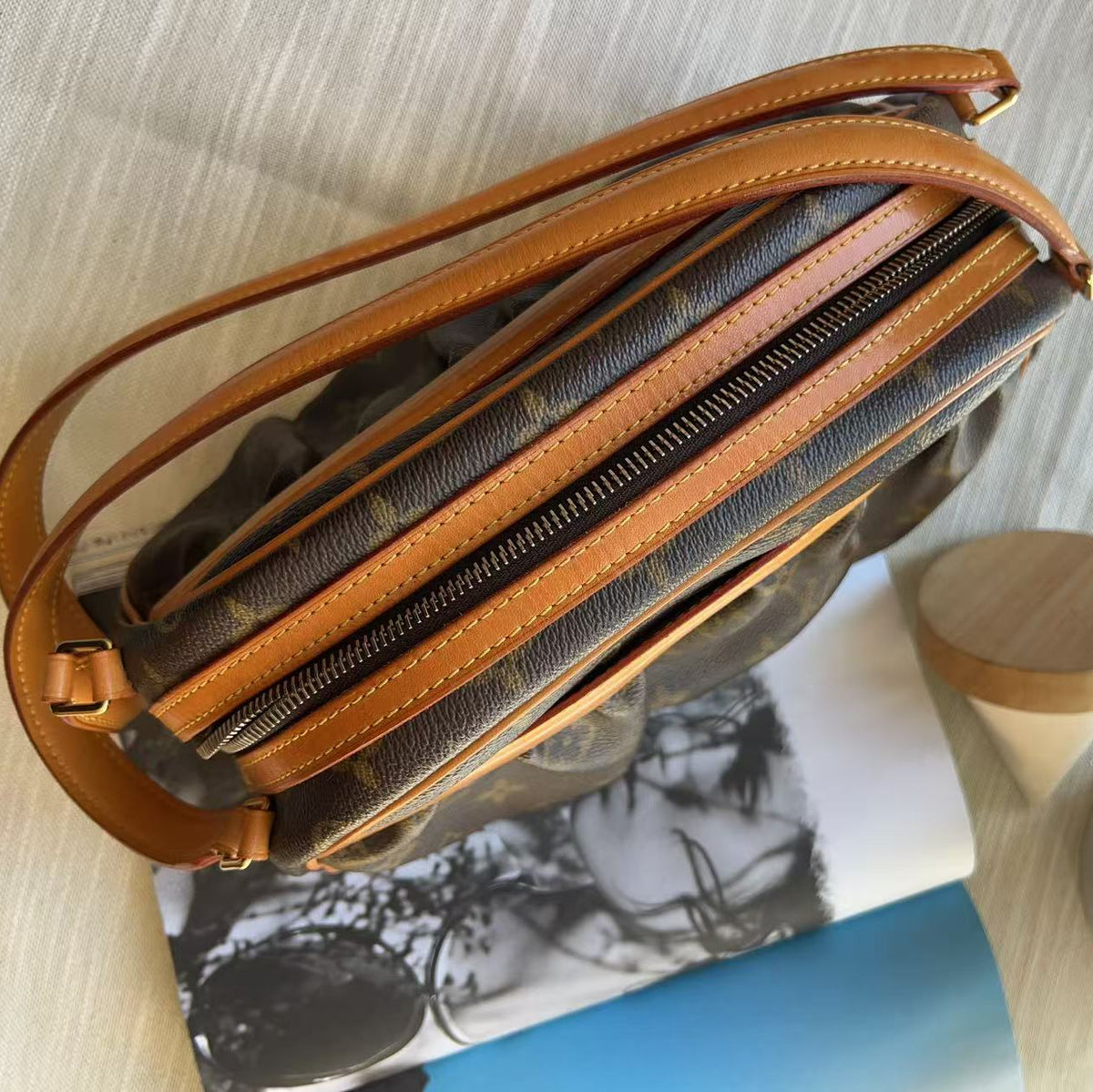 A Louis Vuitton monogram canvas shoulder bag in brown and tan leather, shown from an overhead angle resting on a magazine over a light-colored surface.