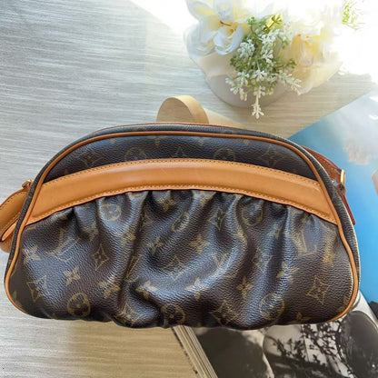 A Louis Vuitton monogram canvas shoulder bag with tan leather trim, shown from a top-down angle on a light-colored surface next to a book and flowers.