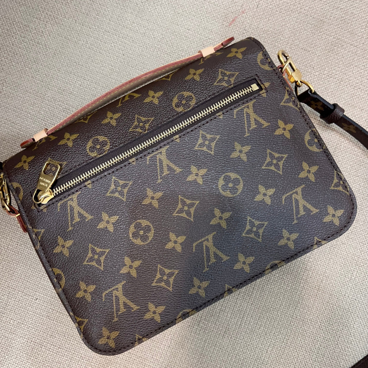 A Louis Vuitton monogram crossbody bag in brown coated canvas with gold-tone hardware, photographed on a light beige textured fabric background.