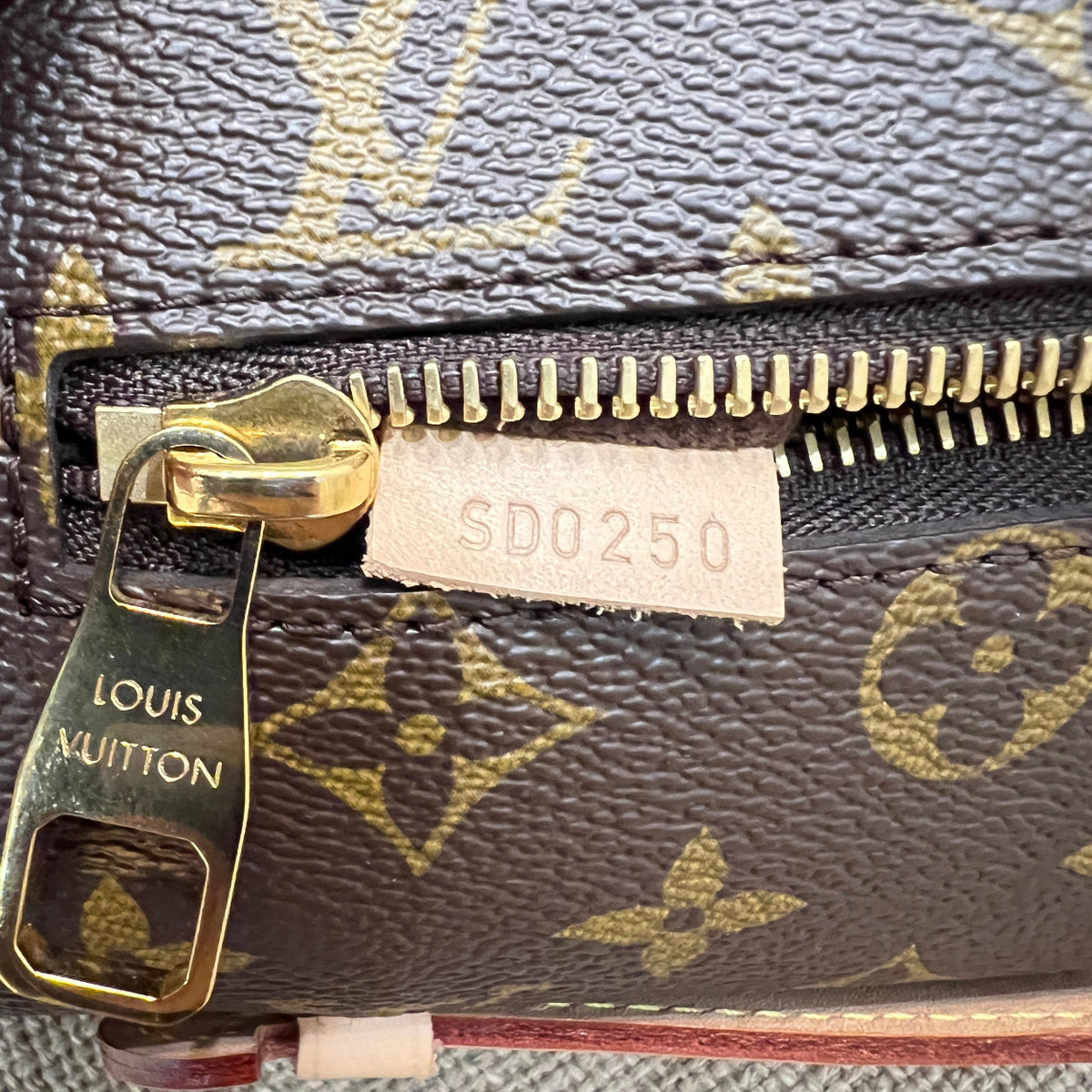 Close-up of a Louis Vuitton monogram zipper pouch showing the dark brown coated canvas with gold monogram print, a gold-tone zipper, and a white paper tag with the code SD0250.