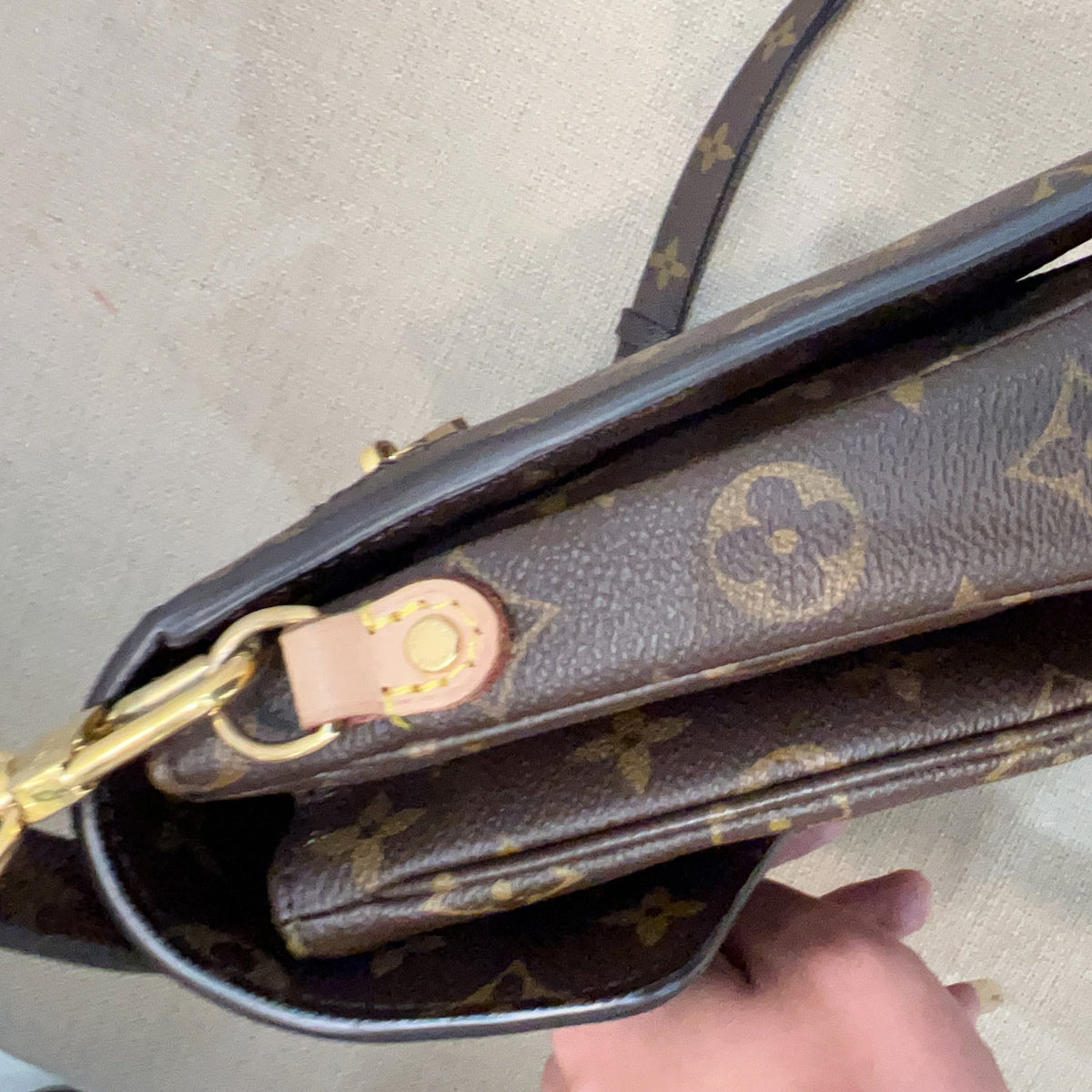 A close-up of a Louis Vuitton monogram shoulder bag in brown coated canvas with gold-tone hardware and a light tan leather detail, held by a hand against a neutral beige background.
