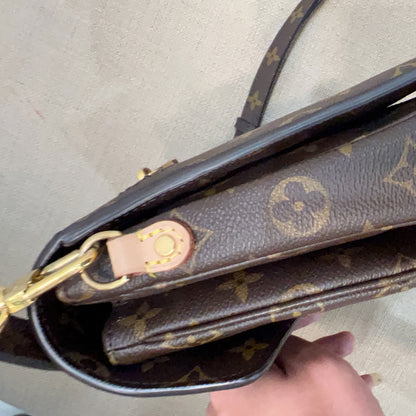 A close-up of a Louis Vuitton monogram shoulder bag in brown coated canvas with gold-tone hardware and a light tan leather detail, held by a hand against a neutral beige background.