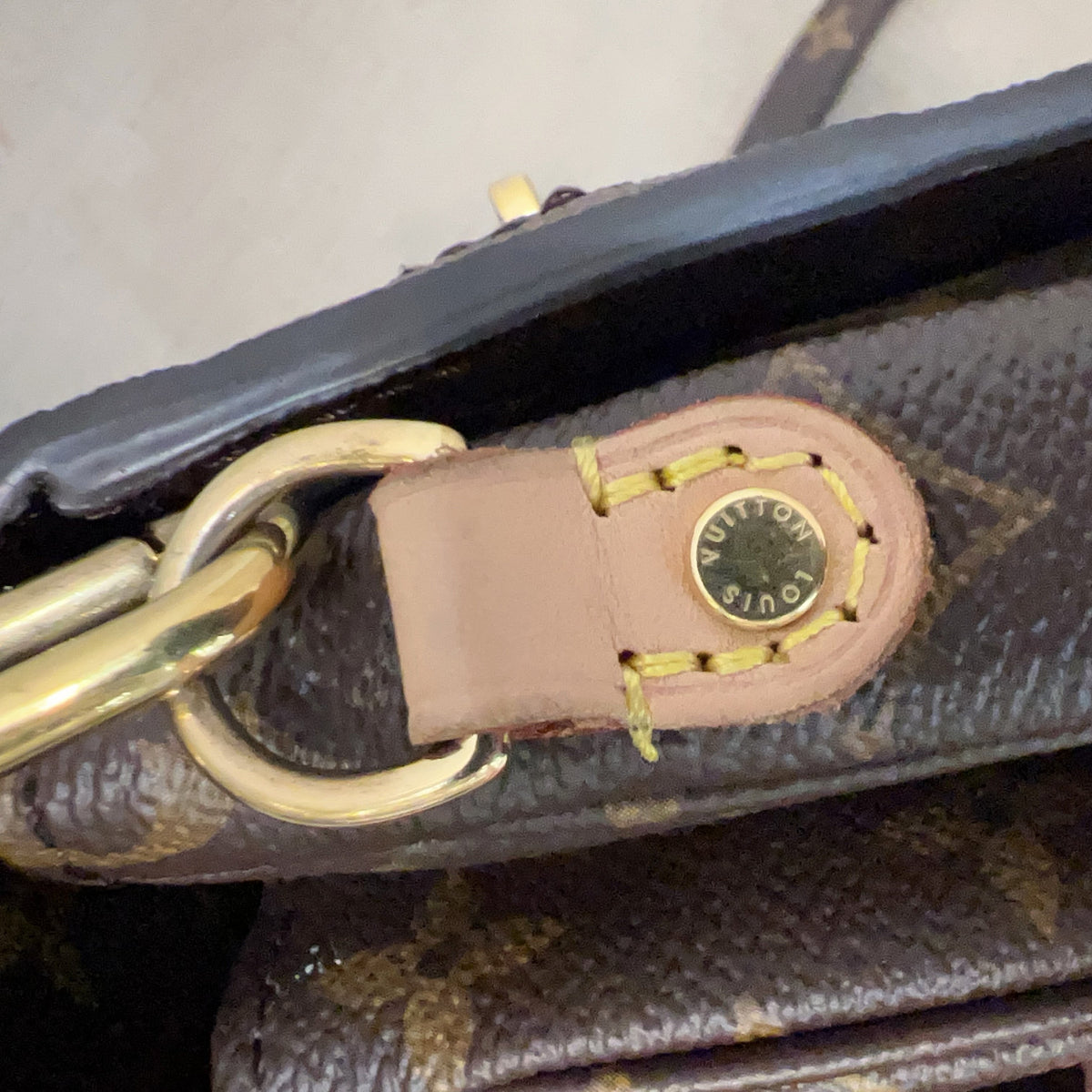 Close-up detail of a Louis Vuitton handbag showing the brown monogram canvas, a tan leather tab, gold-tone hardware, and the circular Louis Vuitton logo stamp.