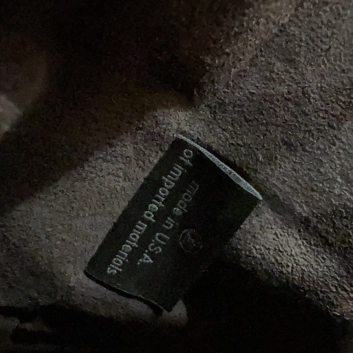 Close-up of a Louis Vuitton interior label stitched onto a dark brown fabric surface, with part of the classic monogram canvas visible at the top right.