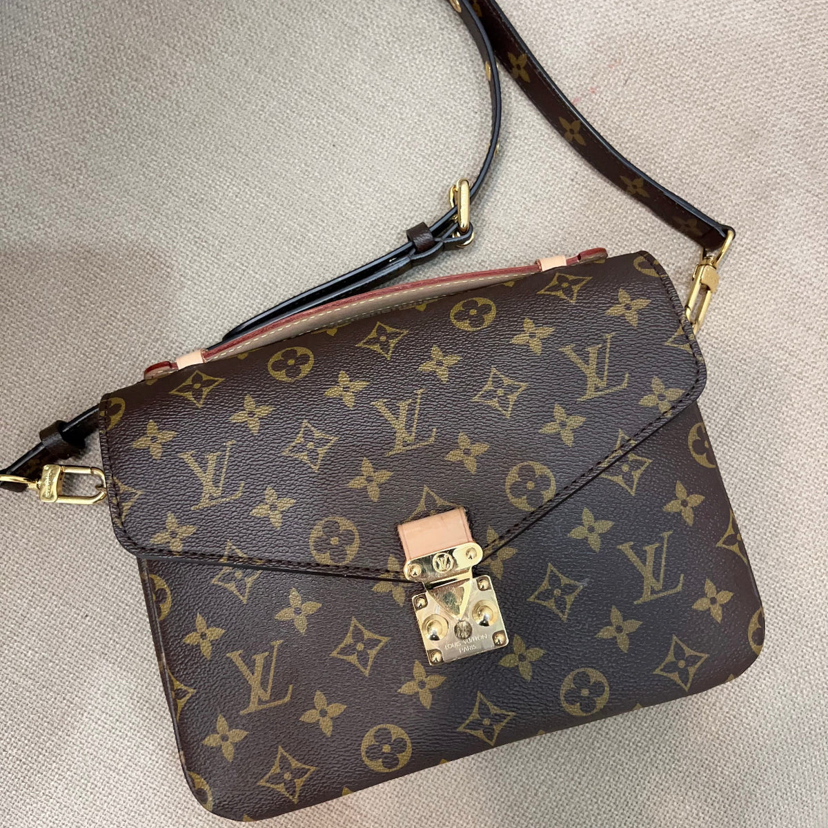 A Louis Vuitton monogram shoulder bag in brown coated canvas with gold-tone hardware, resting on a light beige textured fabric surface.