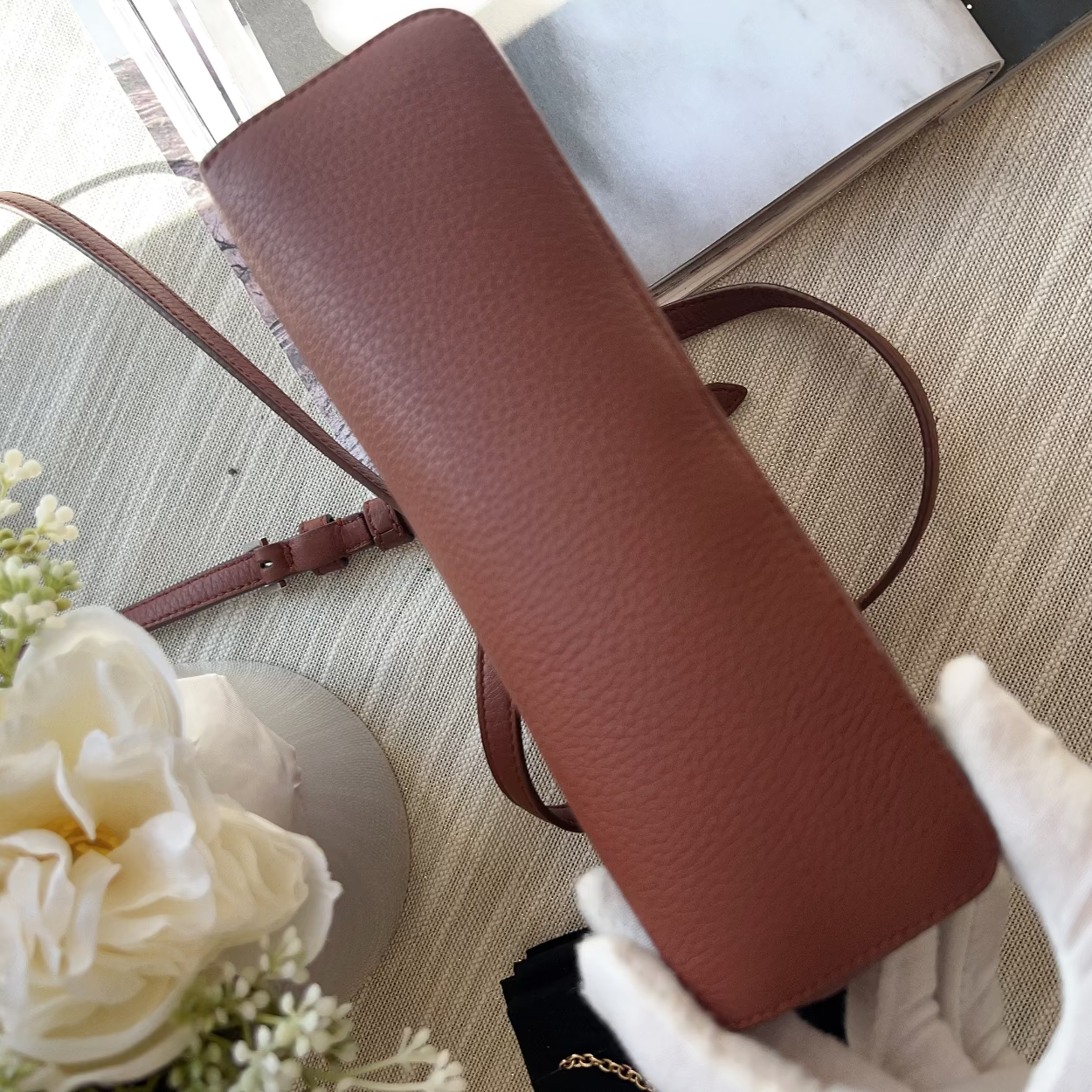 Brown leather handbag with a strap held by a gloved hand on a textured surface with flowers.