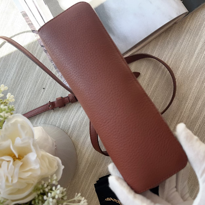 Brown leather handbag with a strap held by a gloved hand on a textured surface with flowers.