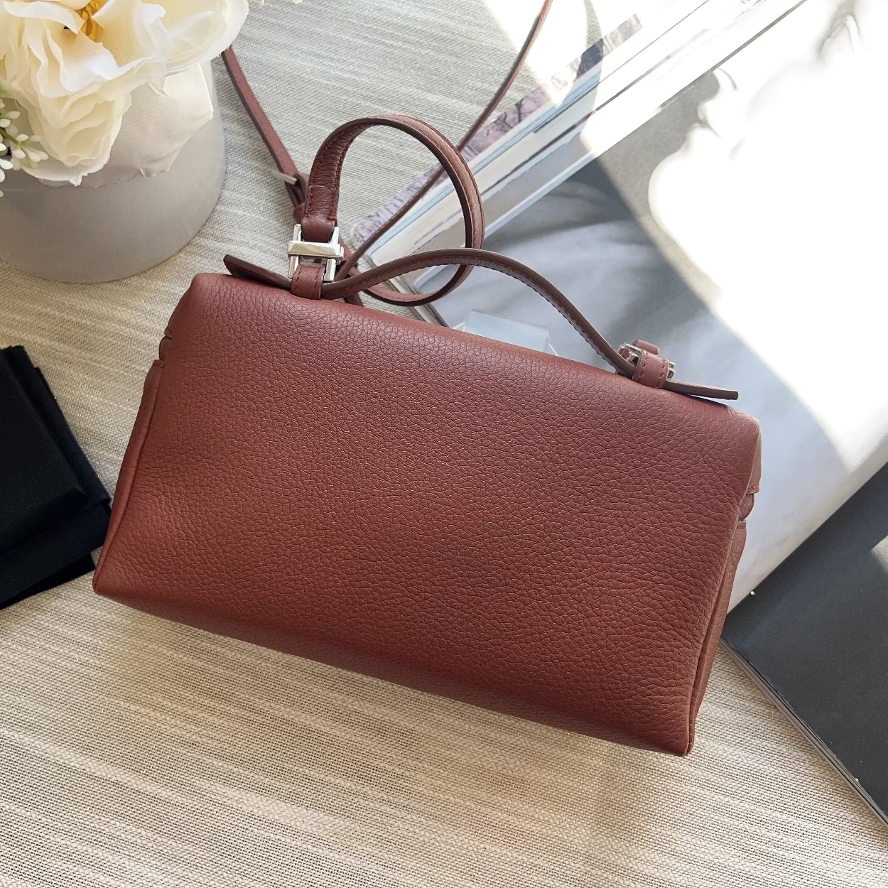 Brown leather handbag on a light wooden surface with a vase of flowers and jewelry box in the background.