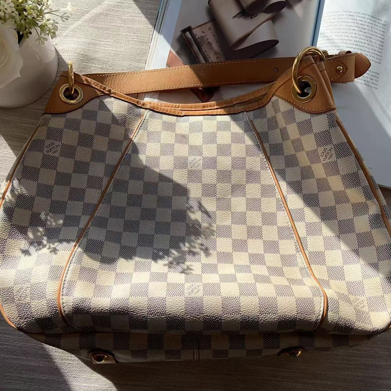 Checkered handbag with brown leather strap on a wooden surface with flowers and an open book in the background