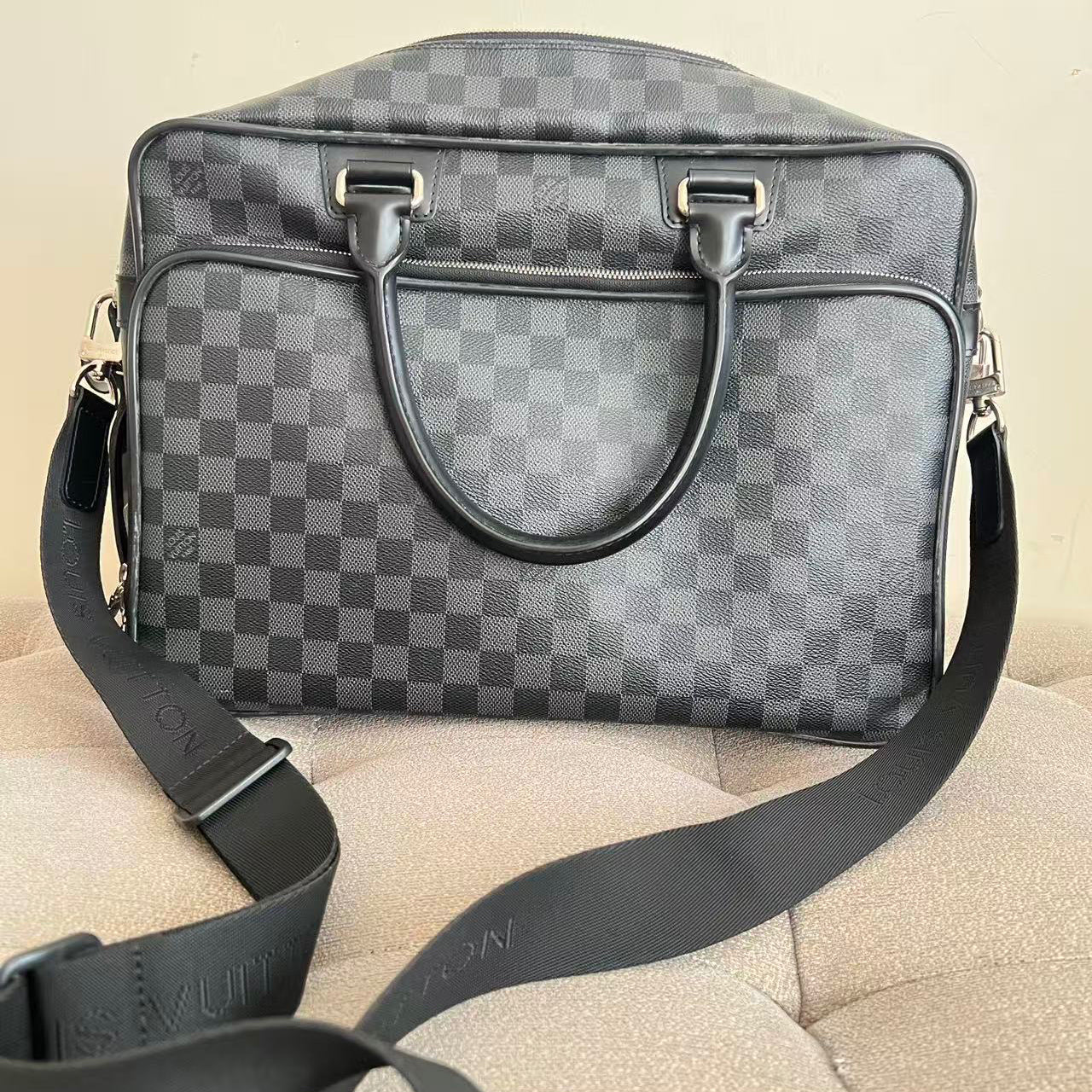 A Louis Vuitton shoulder bag in the signature Damier Graphite checkerboard pattern, featuring dark gray leather handles and a matching adjustable shoulder strap resting on a beige textured surface.