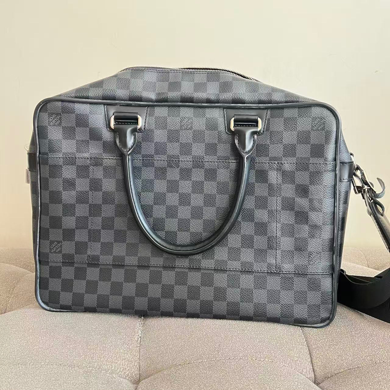 A gray and black checkered Louis Vuitton duffel bag with dark gray leather handles and silver-tone hardware, resting on a beige upholstered surface against a plain beige wall.
