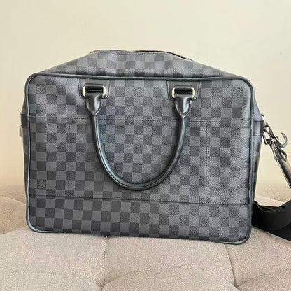 A gray and black checkered Louis Vuitton duffel bag with dark gray leather handles and silver-tone hardware, resting on a beige upholstered surface against a plain beige wall.