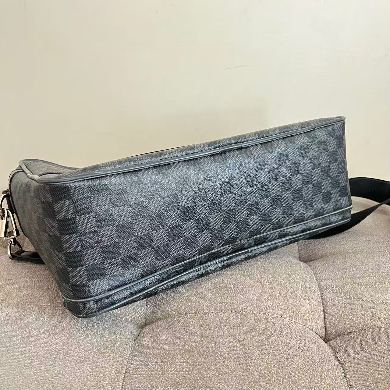 A Louis Vuitton shoulder bag in the Damier Graphite canvas, featuring a dark gray and light gray checkerboard pattern, resting on a beige tufted sofa.
