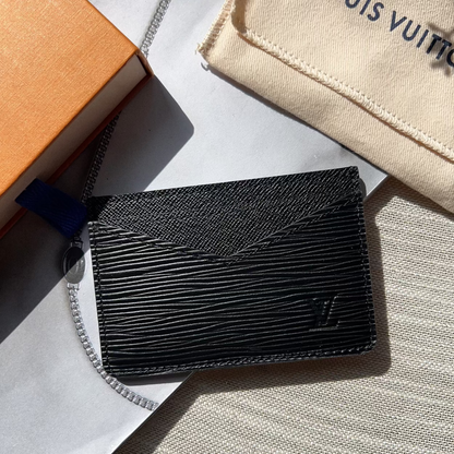 Black textured wallet on a white surface with a beige Louis Vuitton pouch in the background.