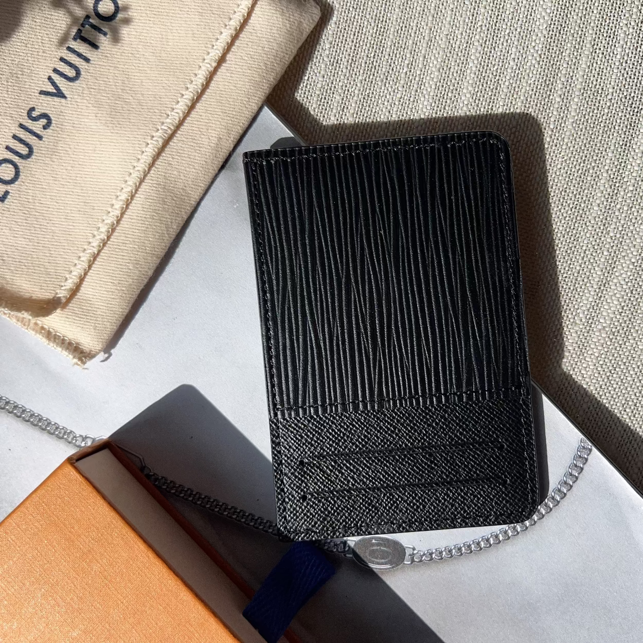 Black textured wallet on a textured surface with a Louis Vuitton pouch in the background.