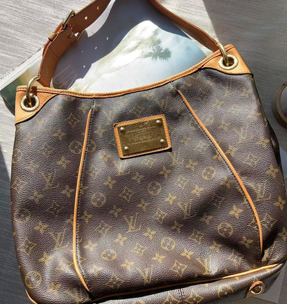 A Louis Vuitton monogram shoulder bag in brown coated canvas with tan leather trim and gold-tone hardware, photographed on a light textured surface with sunlight and shadows.