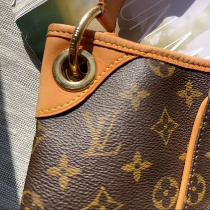 Close-up of a Louis Vuitton monogram canvas bag in brown with tan leather trim and a gold-tone metal grommet, resting on a textured gray surface.