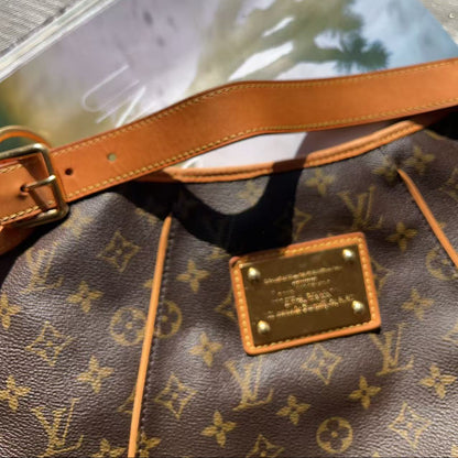 A close-up of a Louis Vuitton monogram canvas bag with tan leather trim and a gold-tone logo plaque, resting on a gray textured surface with a magazine partially visible underneath.