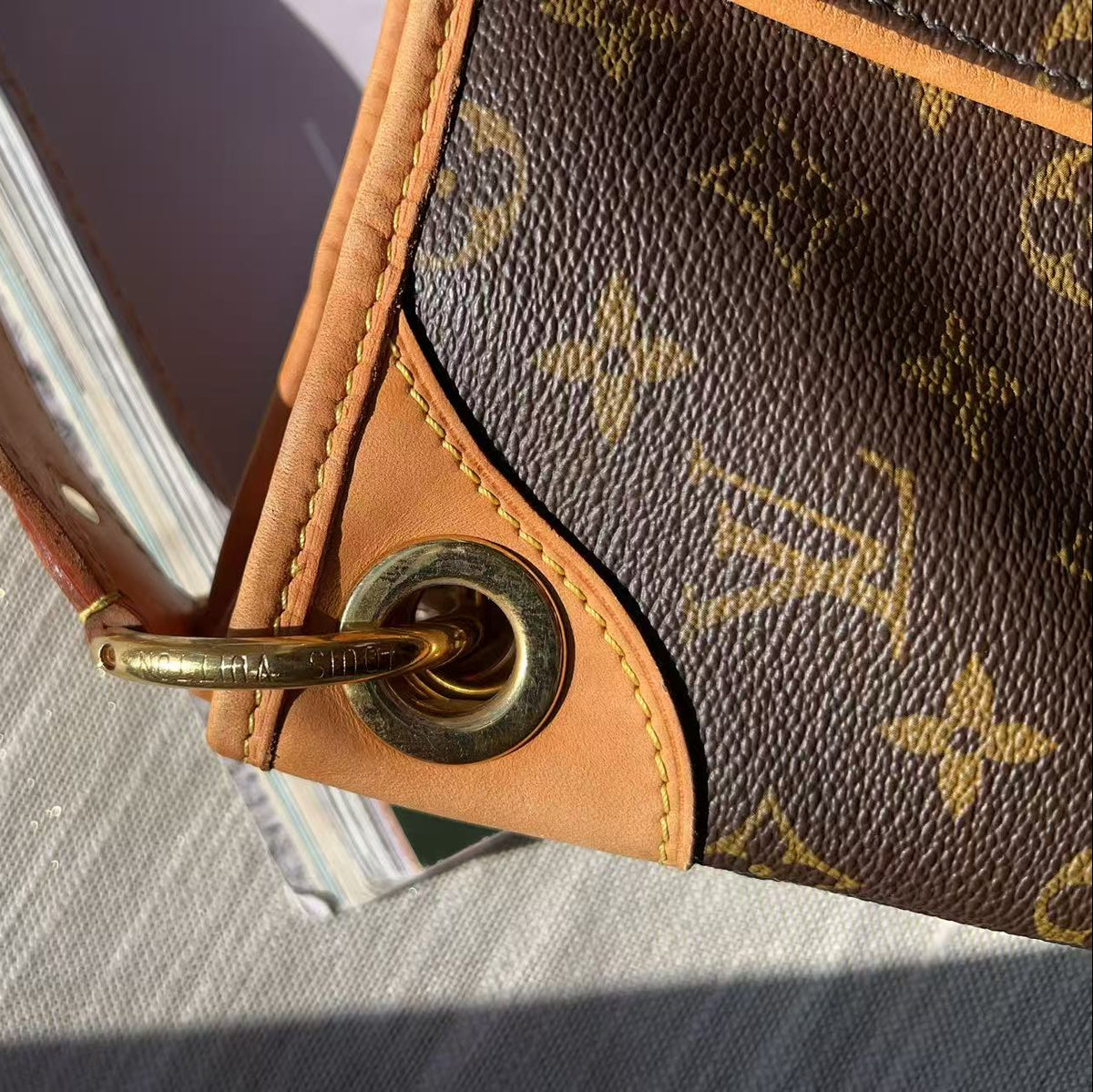 Close-up of a Louis Vuitton monogram handbag corner with tan leather trim, gold-tone metal grommet, and part of the signature brown-and-gold monogram canvas on a light gray textured surface.