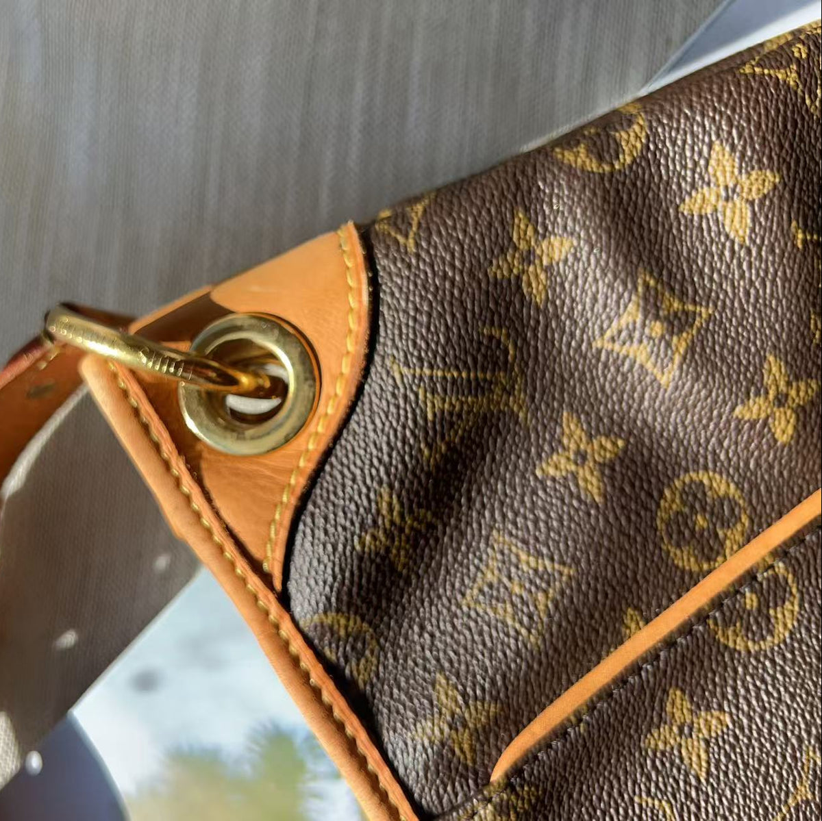 Close-up of a Louis Vuitton shoulder bag corner showing the brown monogram canvas, tan leather trim, and gold-tone metal grommet and strap attachment.