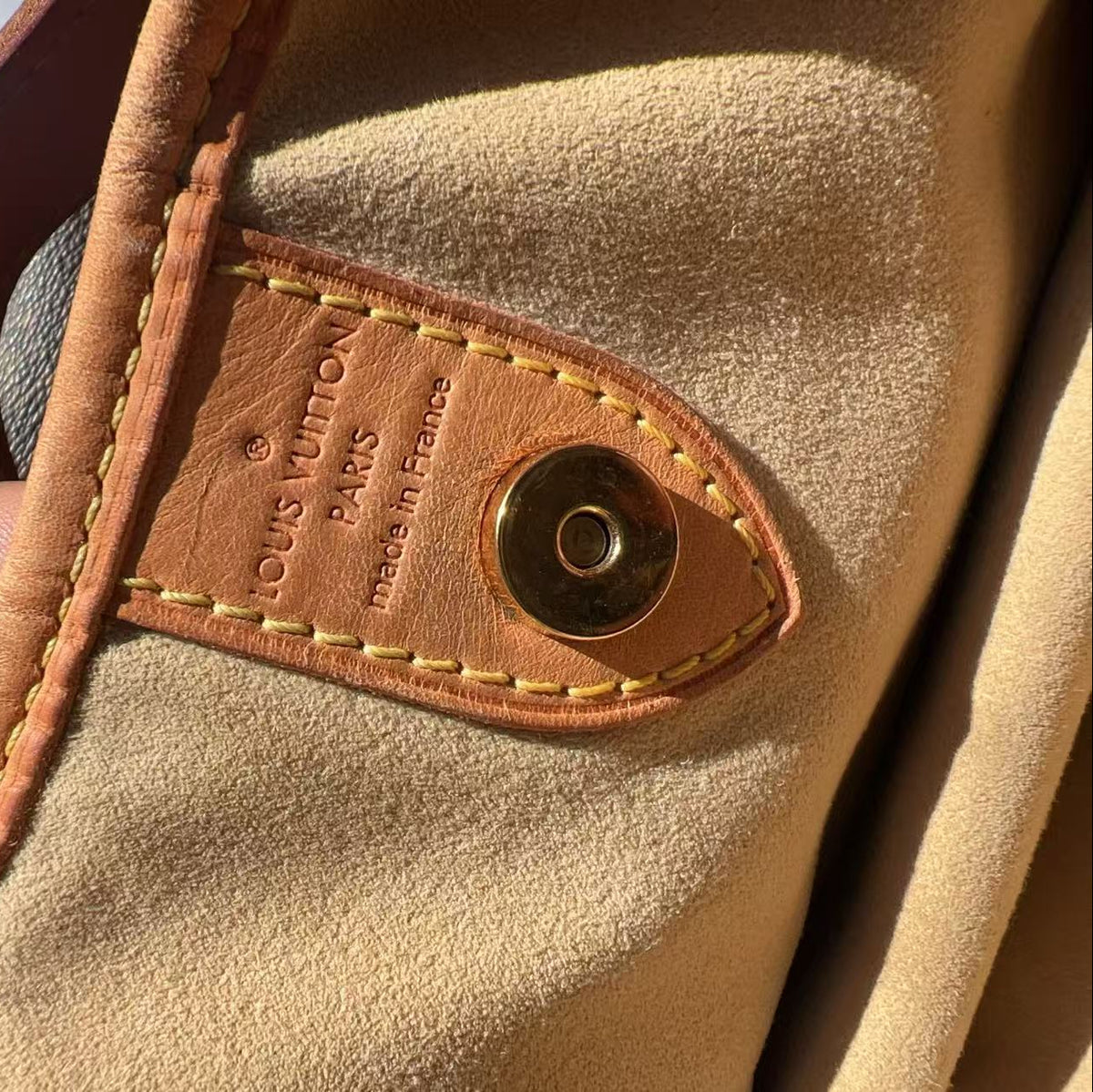 Close-up of a Louis Vuitton bag interior showing a tan leather tab stamped 'LOUIS VUITTON PARIS made in France' with a brass snap button, attached to a beige suede or nubuck lining, held by a hand.