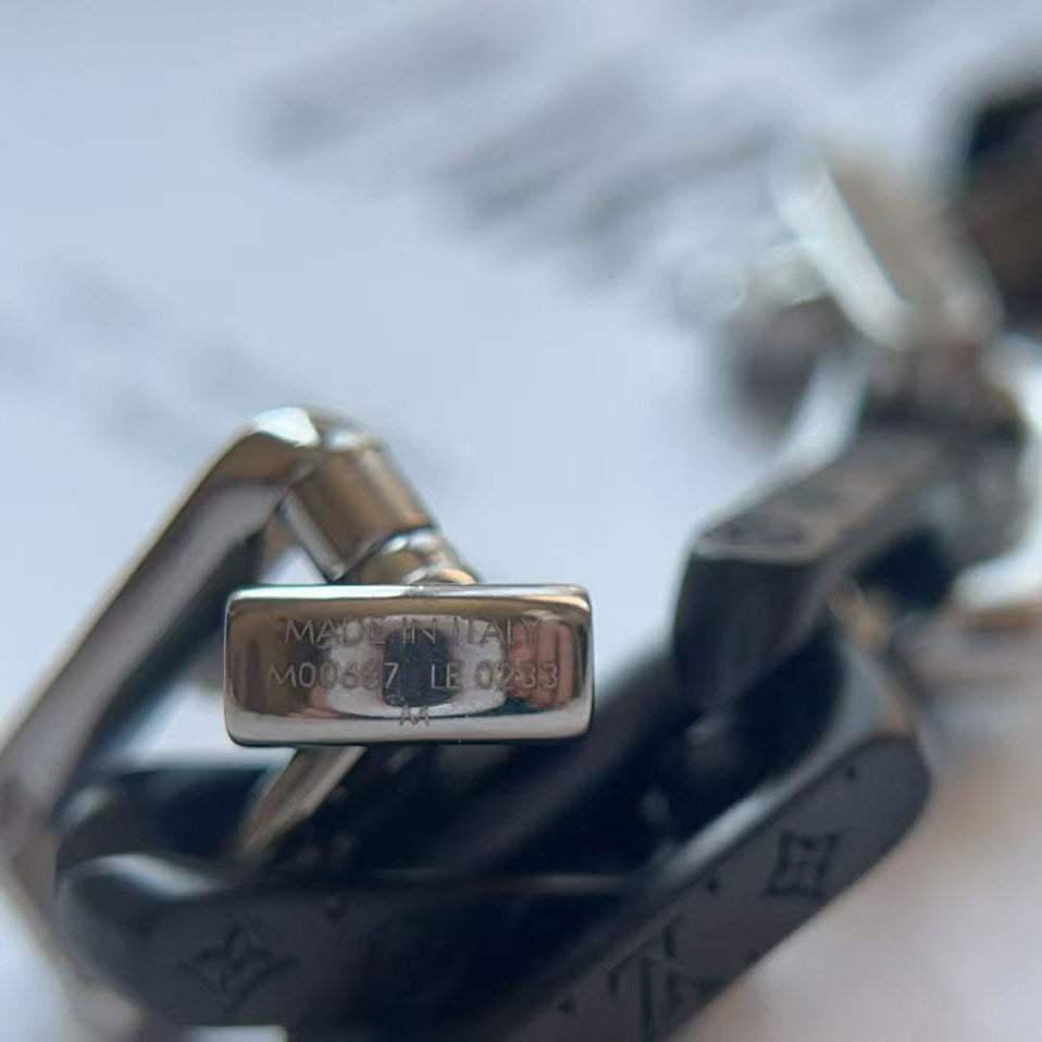 Close-up of a silver-toned metal buckle and chain with engraved text on a black monogram-patterned strap, photographed on a light blurred background.