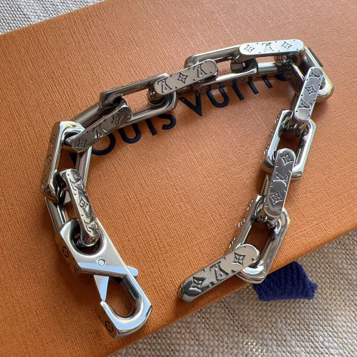 A silver-tone chain bracelet with engraved Louis Vuitton monogram and LV motifs, resting on an orange Louis Vuitton box.