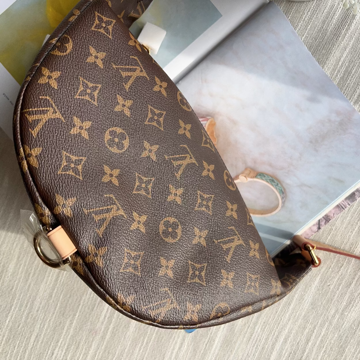 A Louis Vuitton monogram shoulder bag in brown coated canvas with gold-tone hardware, resting on a light-colored textured surface with printed papers underneath.