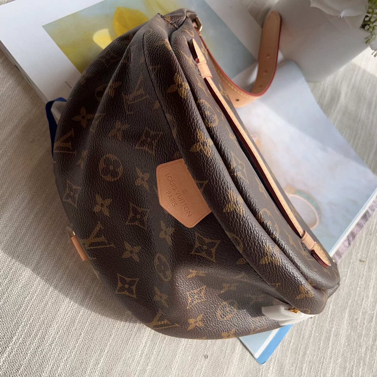 A Louis Vuitton monogram canvas shoulder bag in brown with tan leather trim, resting on a white textured surface with a magazine and flowers in the background.