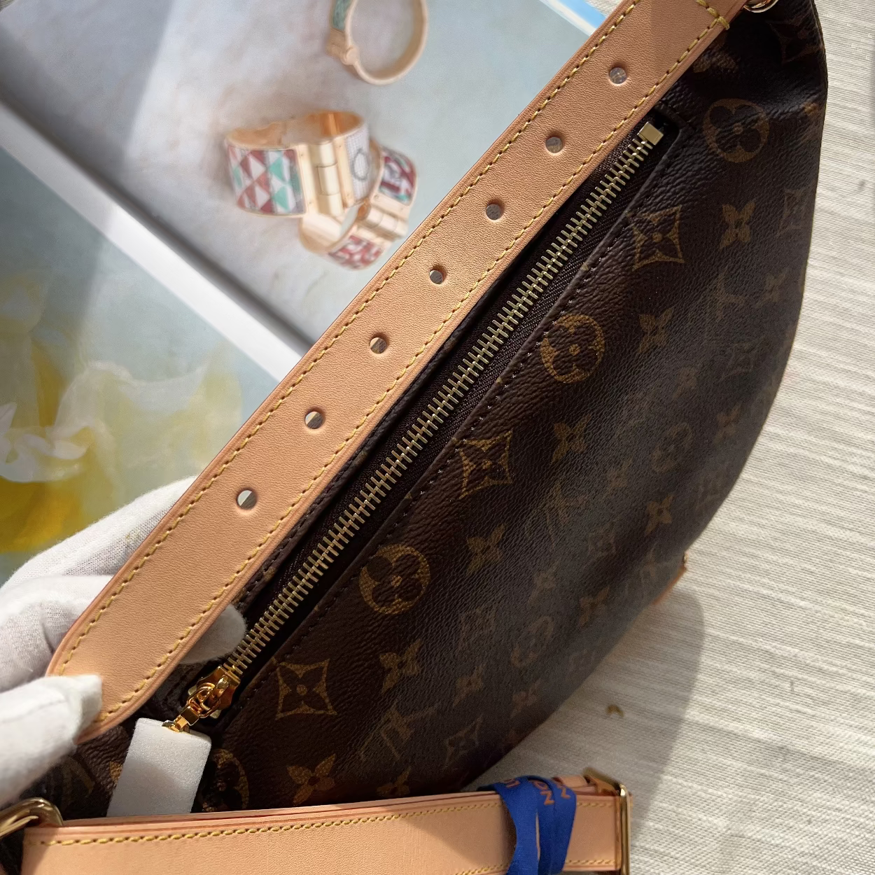 A close-up of a Louis Vuitton monogram shoulder bag in dark brown coated canvas with tan leather trim and gold-tone hardware, photographed on a light wood surface with a magazine in the background.