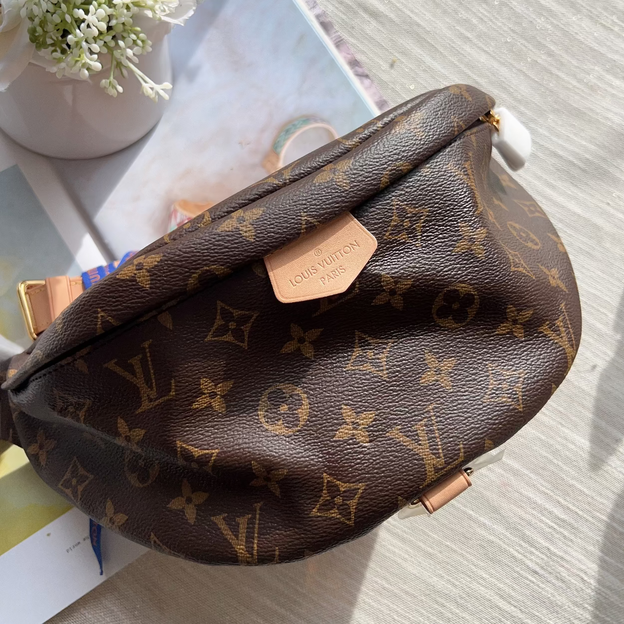 Louis Vuitton monogram canvas shoulder bag in brown with gold-tone hardware, photographed on a light wood surface with a small vase of white flowers and printed cards nearby.