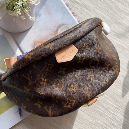 Louis Vuitton monogram canvas shoulder bag in brown with gold-tone hardware, photographed on a light wood surface with a small vase of white flowers and printed cards nearby.