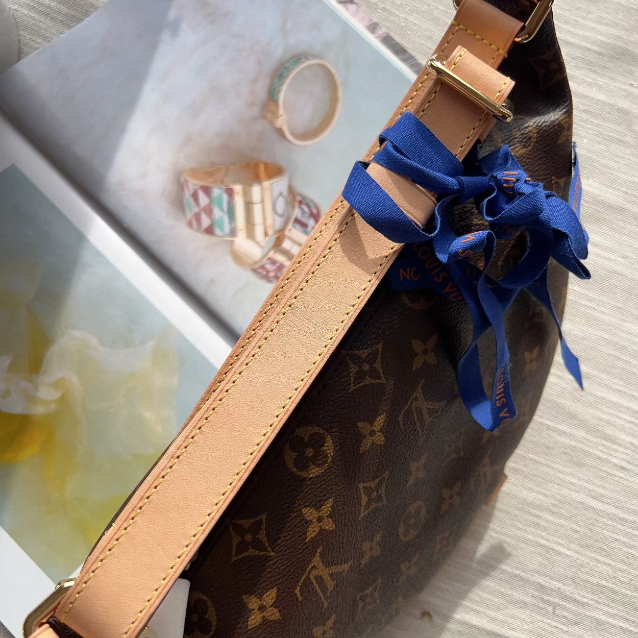 A Louis Vuitton monogram canvas belt bag with tan leather trim and a blue satin ribbon bow, shown resting on a light wood surface next to an open magazine.