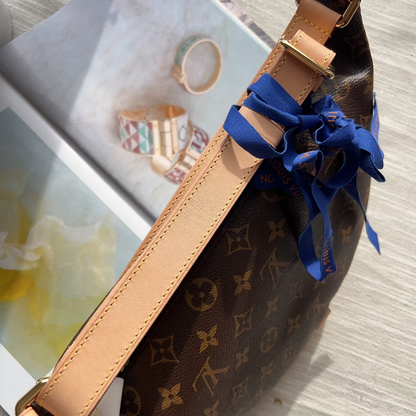 A Louis Vuitton monogram canvas belt bag with tan leather trim and a blue satin ribbon bow, shown resting on a light wood surface next to an open magazine.